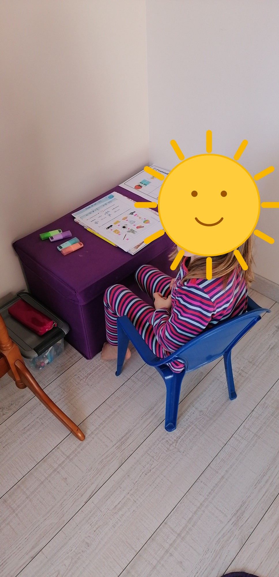 Enfant assise à son bureau, visage masqué par un soleil dessiné, pendant un temps de concentration à la maison