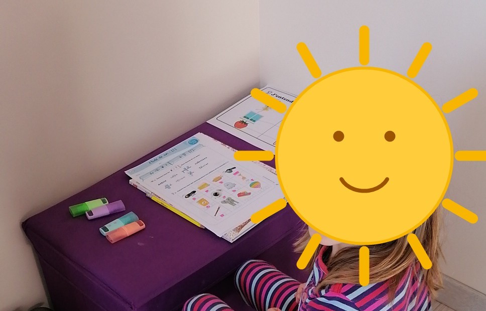 Enfant assise à son bureau, visage masqué par un soleil dessiné, pendant un temps de concentration à la maison